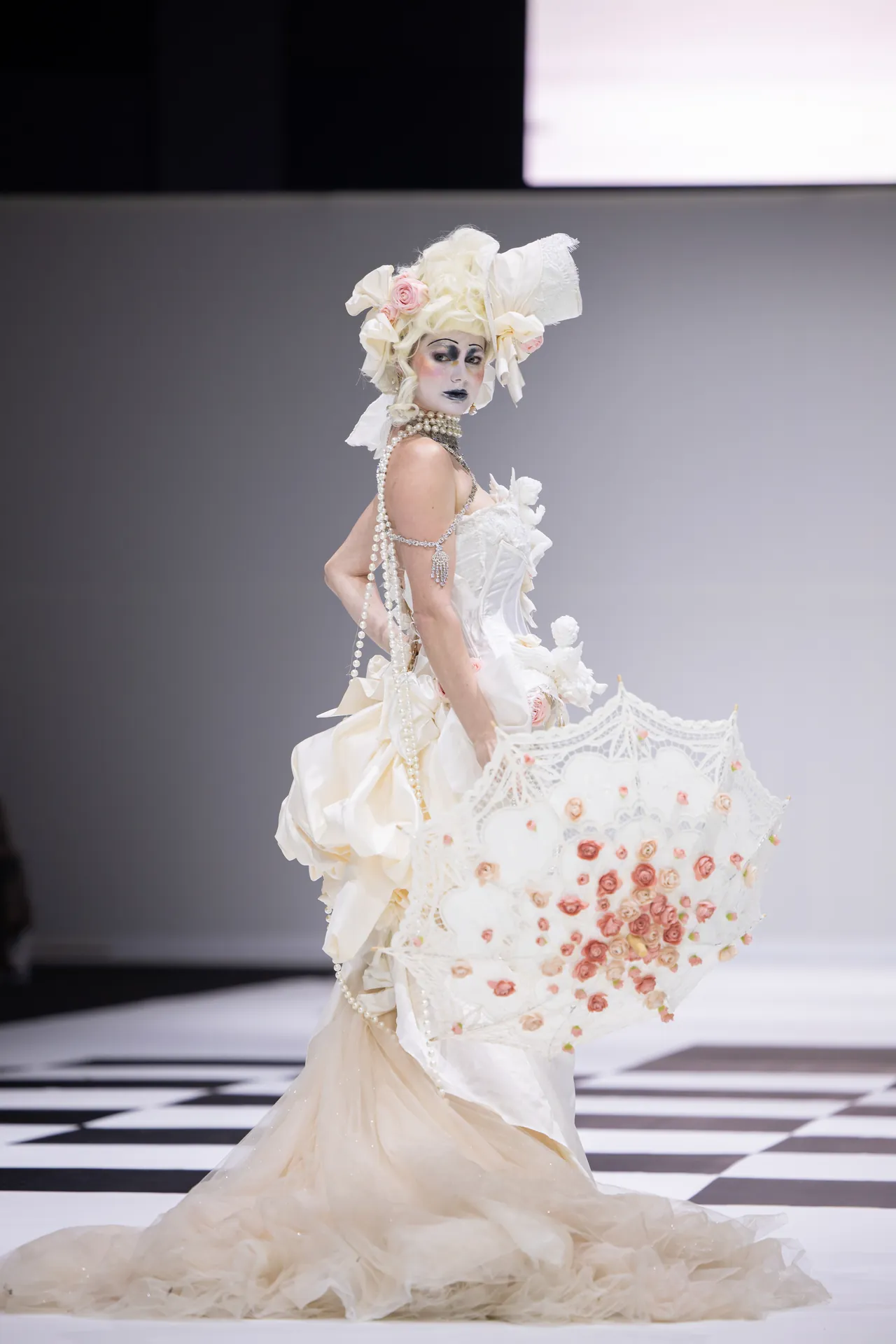 Model on a fashion runway in a voluminous white gown with rose details and a large floral headpiece.