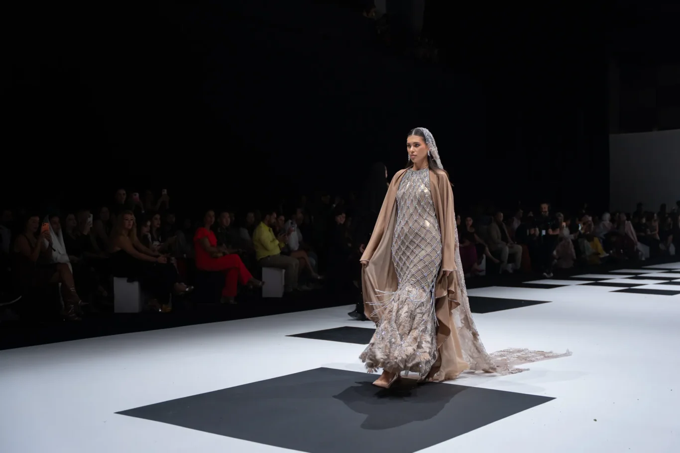 Model walks the runway in a shimmering silver lattice gown with a beige cape, audience seated dimly in the background.