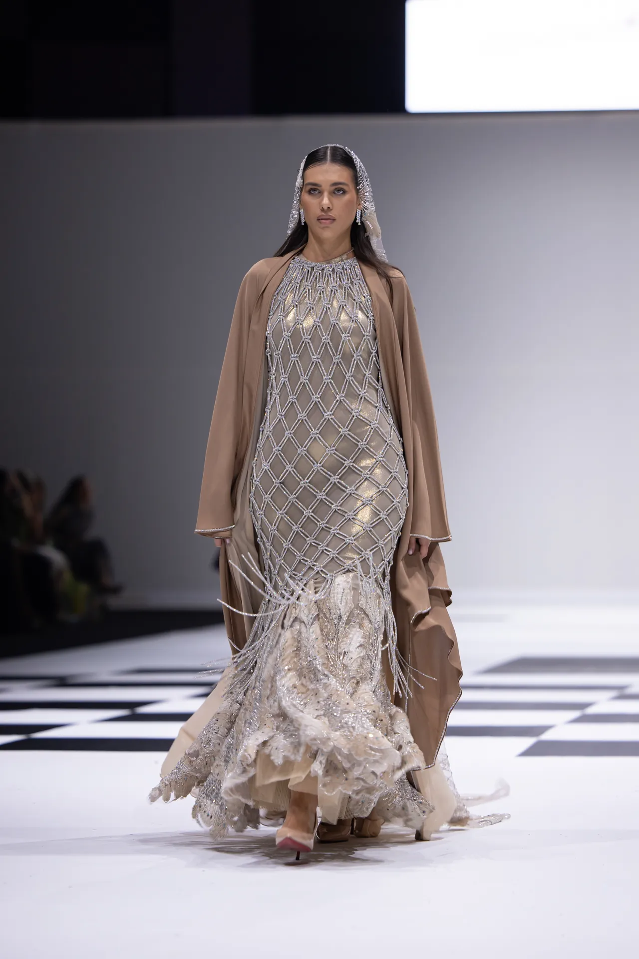 Model on a runway wearing a beige, beaded lattice gown with a sheer veil and flowing cape.