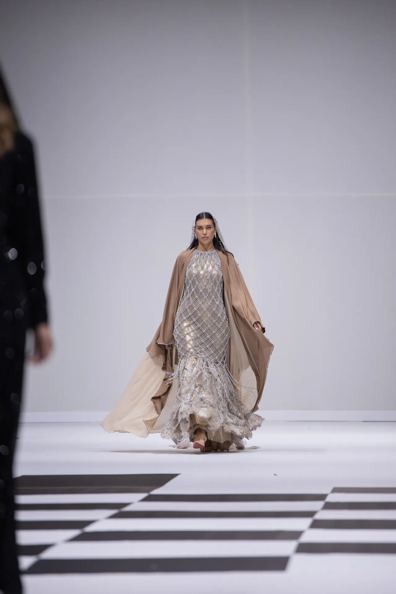 Model walking a fashion runway in a silver, beaded gown with a flowing beige cape.