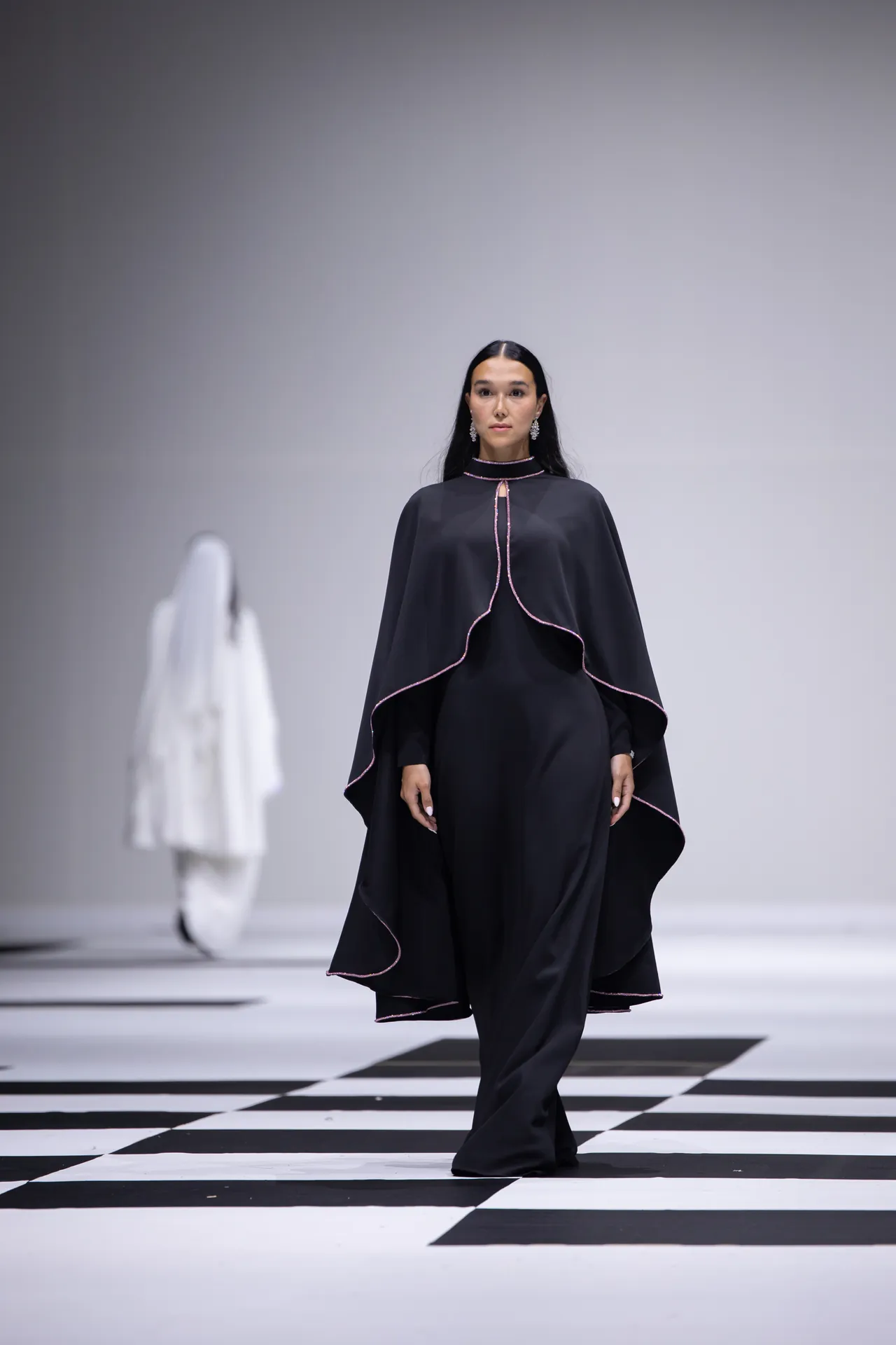 Model walks a fashion runway in a black cape dress with pink piping, standing over a bold black-and-white checkered floor.
