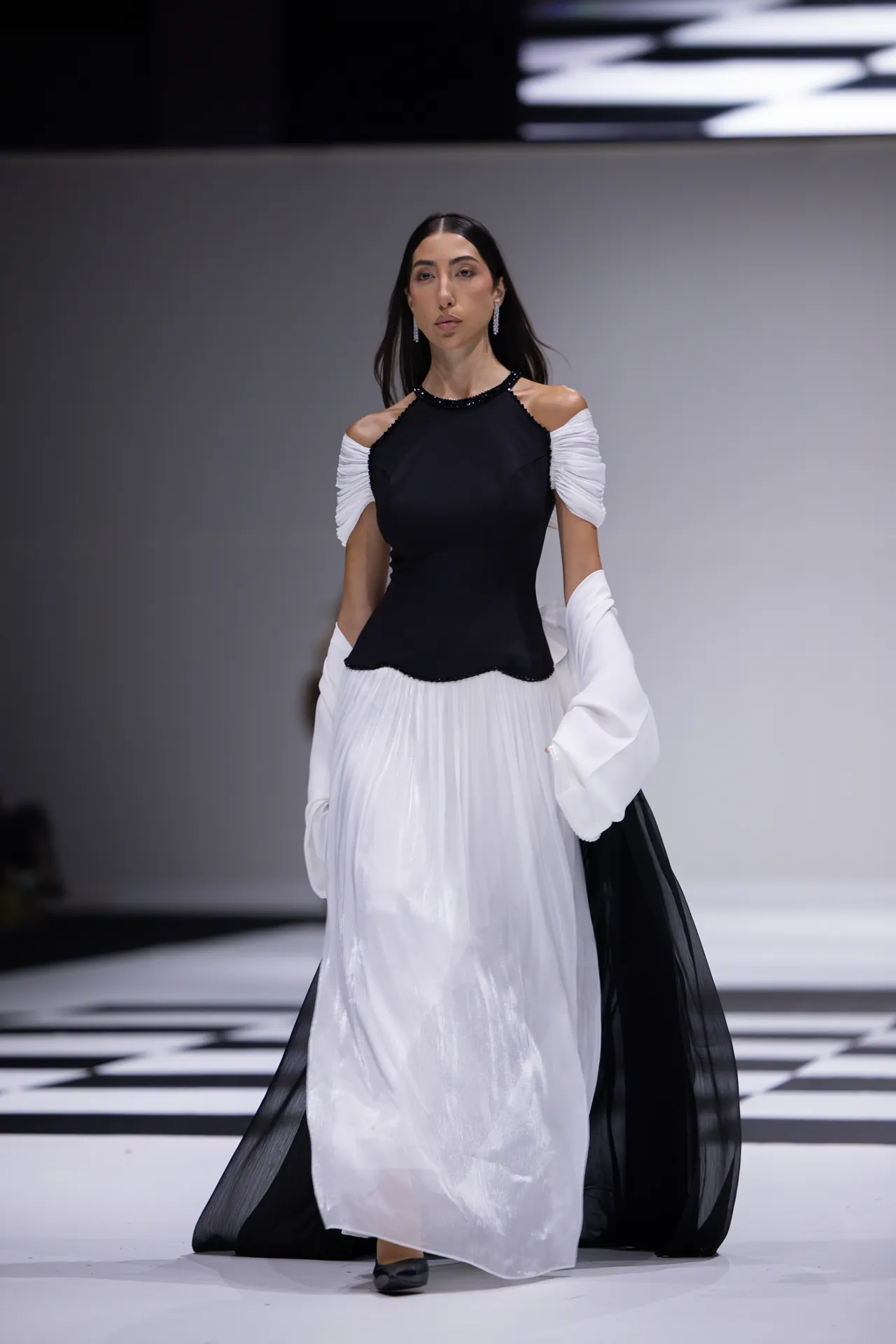 Model walks the runway in a two-tone gown: black bodice with white off-shoulder sleeves and a flowing white skirt with black sheer accents behind her.