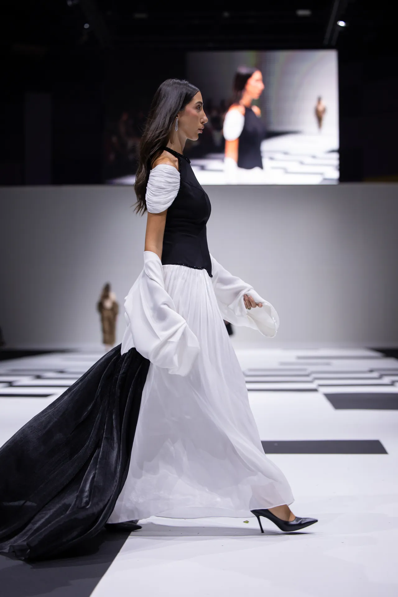 Side view of a female model walking the runway in a black bodice and white skirt with a long black train behind her.