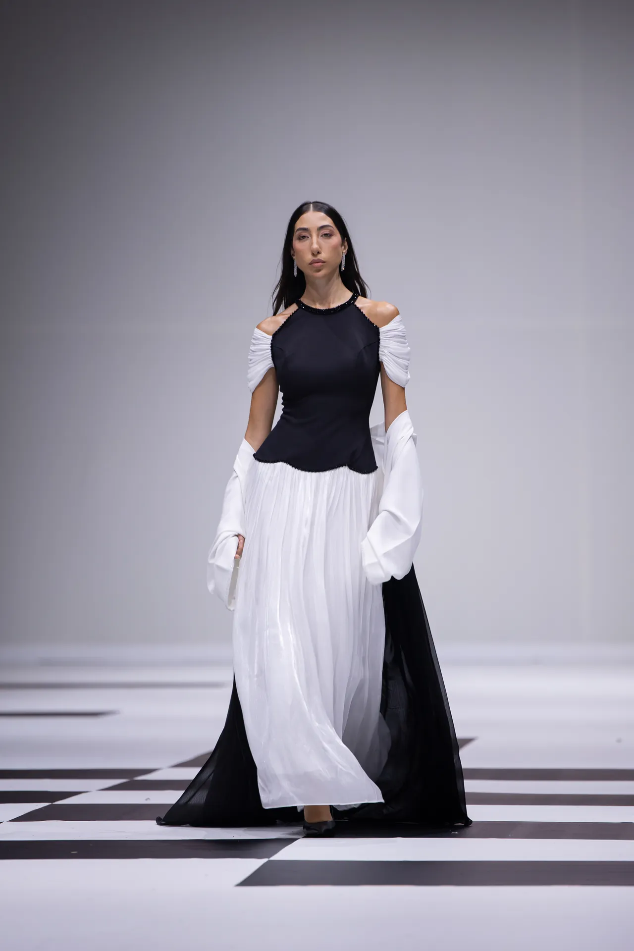 Runway model walks wearing a black bodice with white off-shoulder sleeves and a flowing two-tone skirt on a black-and-white checkerboard runway.