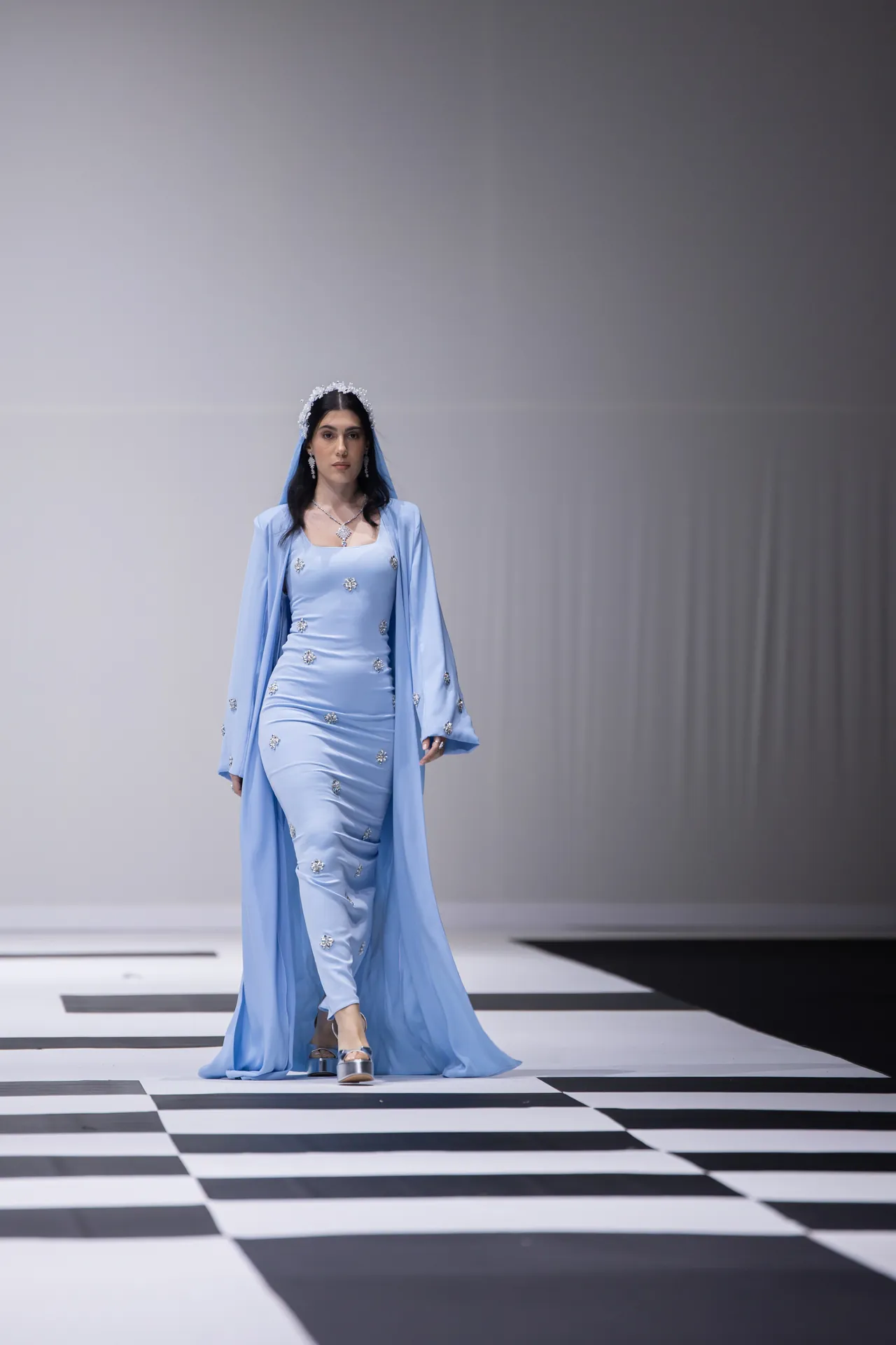 Model walks the runway in a light blue gown with a matching cape and jeweled accessories, on a black-and-white checkerboard catwalk.