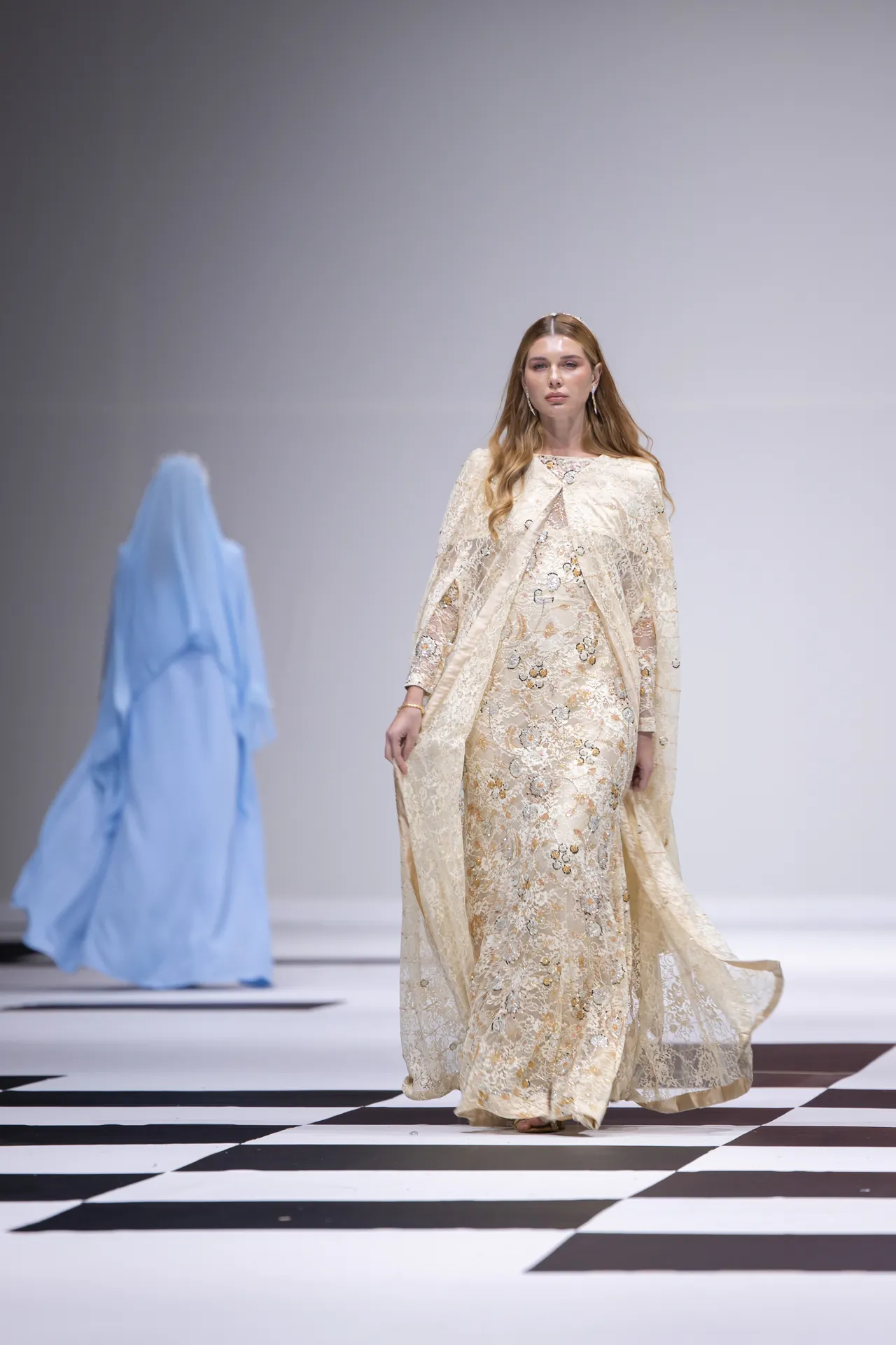 Model walks the runway in a cream lace gown with a matching cape, on a black-and-white checkered floor. Blue fabric backdrop to the left.