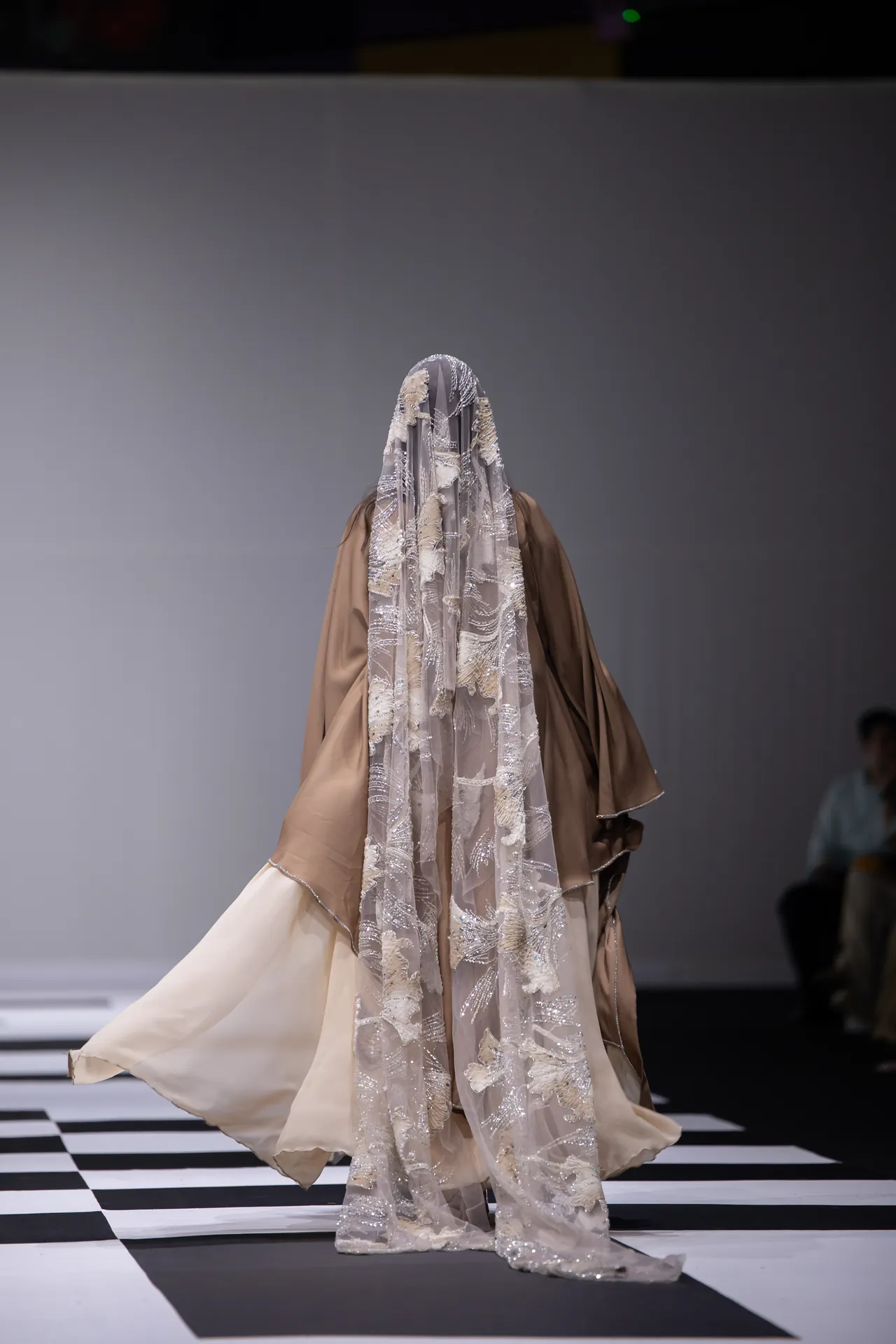 Back view of a model walking a black-and-white checkered runway in a flowing beige gown with an ornate lace veil covering her head and shoulders.