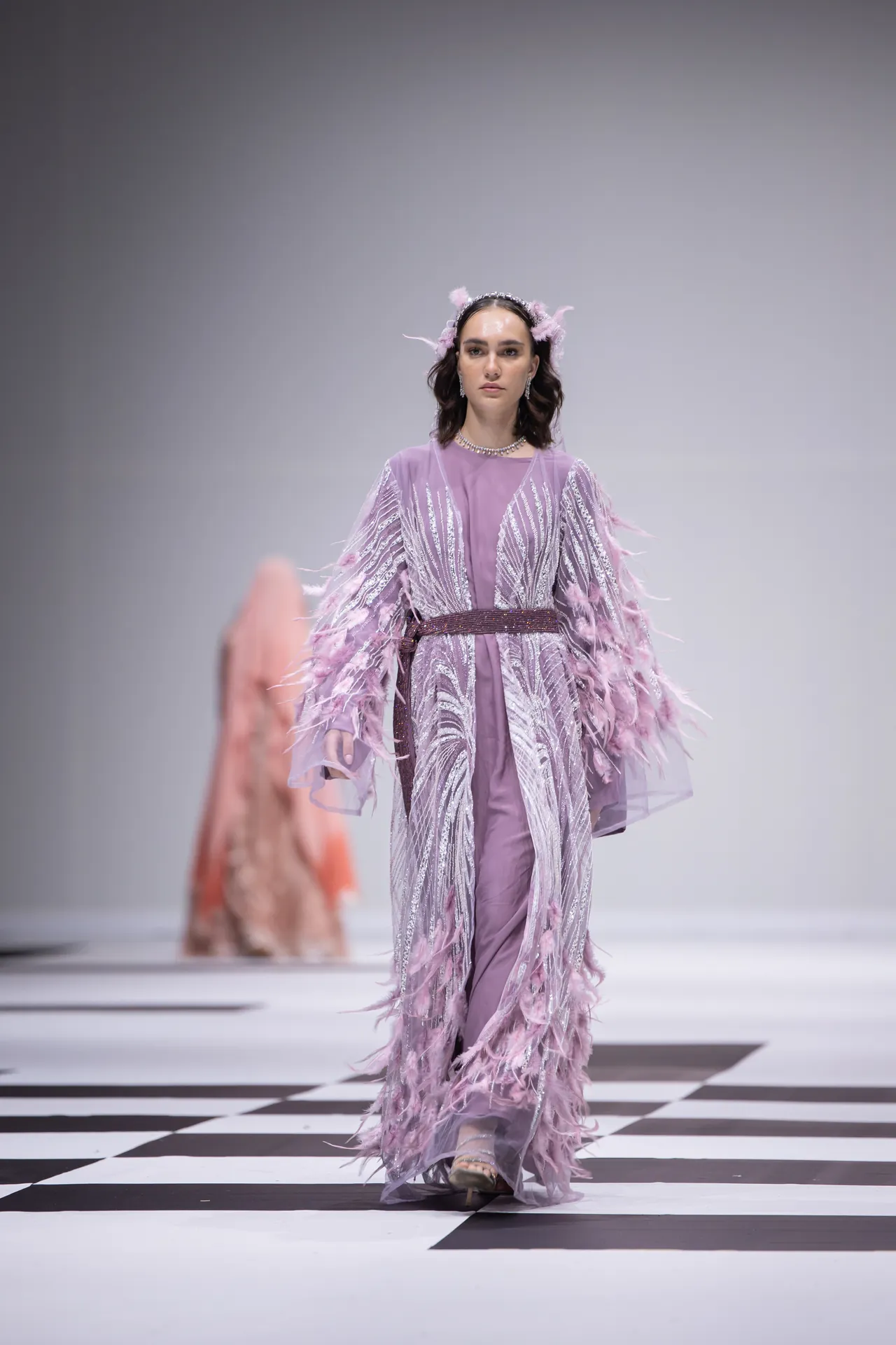 Model walks the runway in a lavender gown with silver fringe and feather accents on a black-and-white checkered runway.