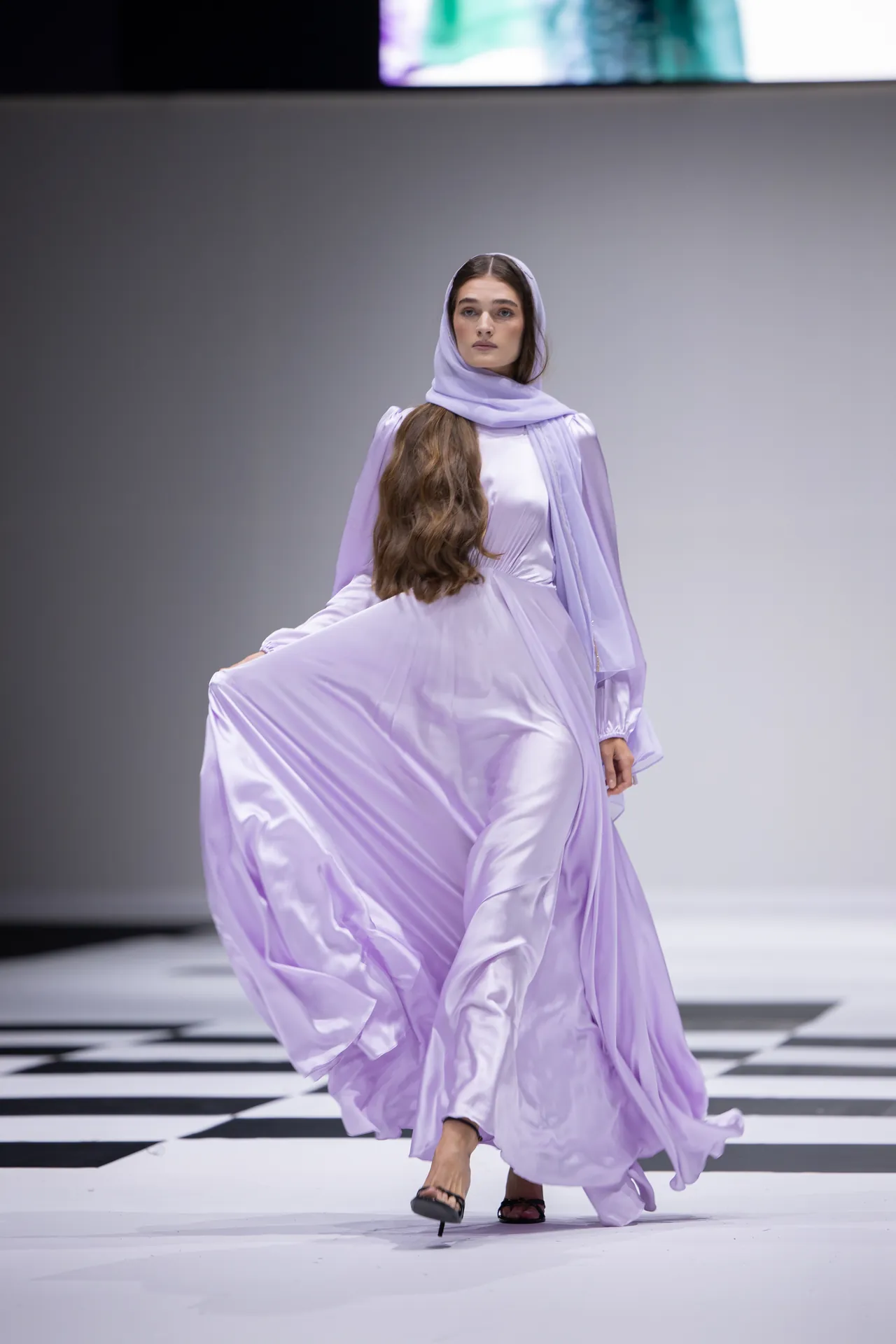 Model in a flowing lavender dress and matching headscarf walks down a black-and-white checkerboard runway.