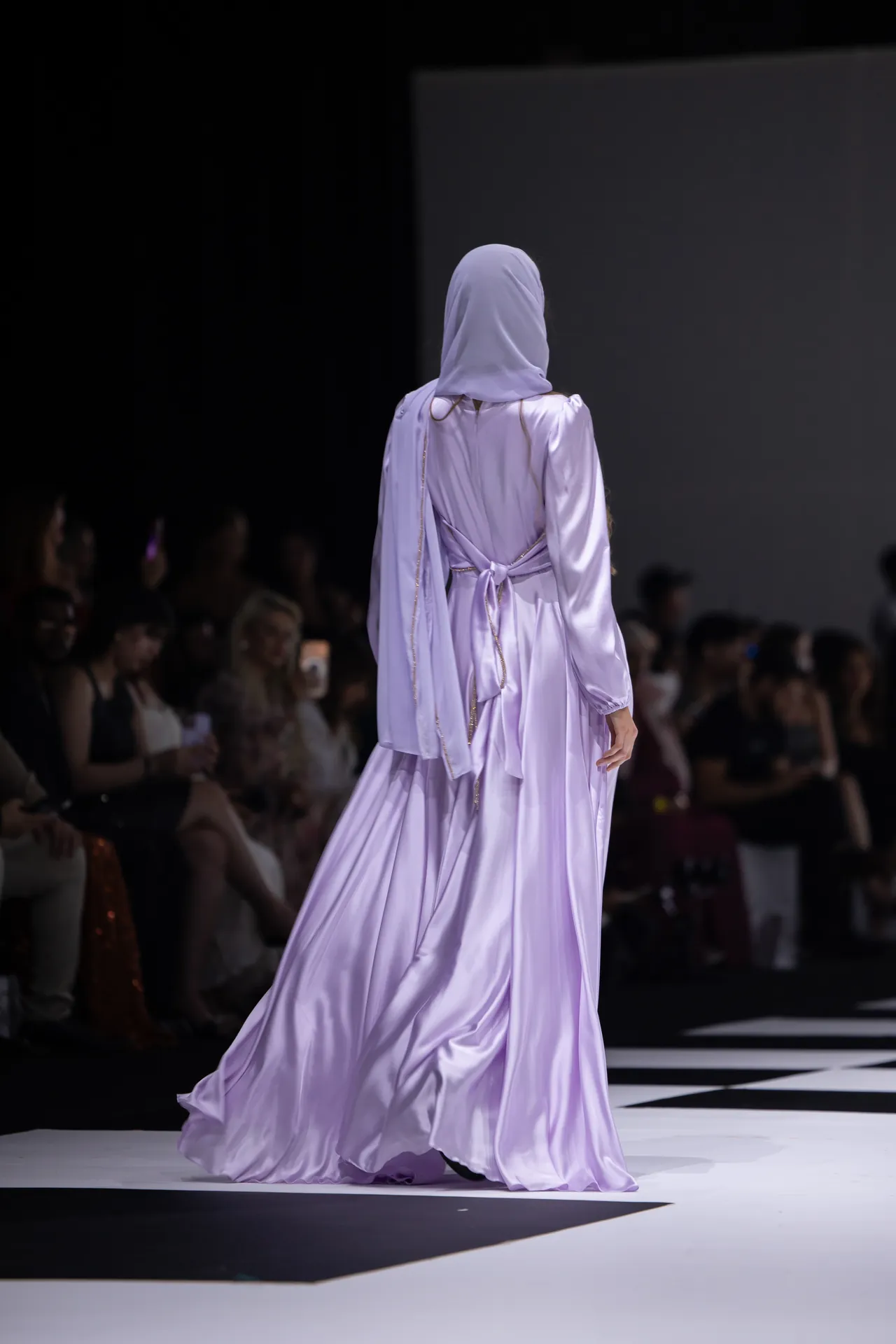 Model walking the runway in a lavender hijab and flowing satin gown, shown from the back with audience in the background.