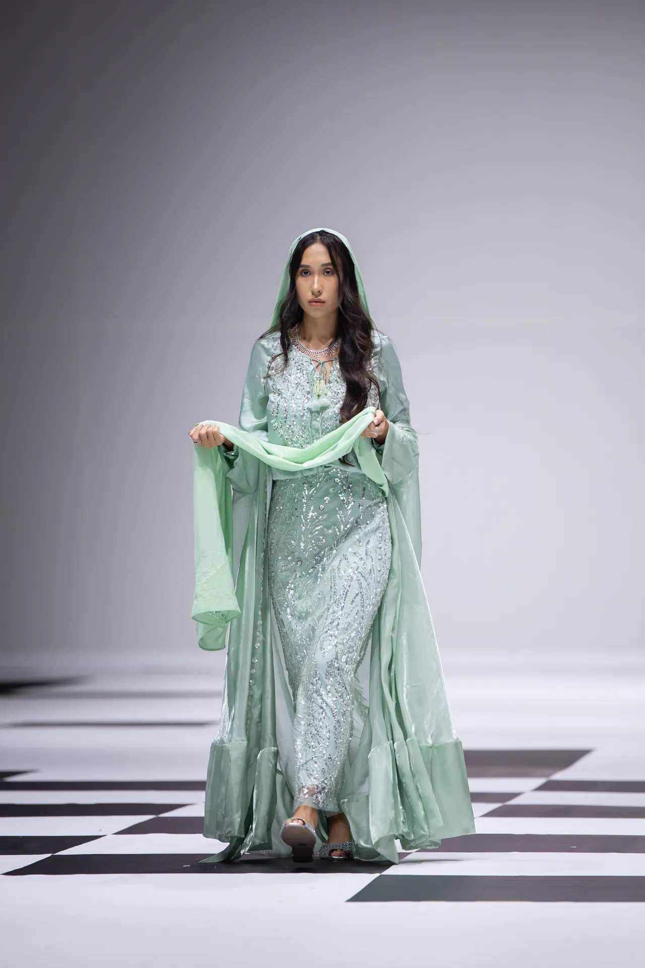 Model walking a fashion runway in a mint green embroidered gown with matching scarf, on a black-and-white checkered catwalk.