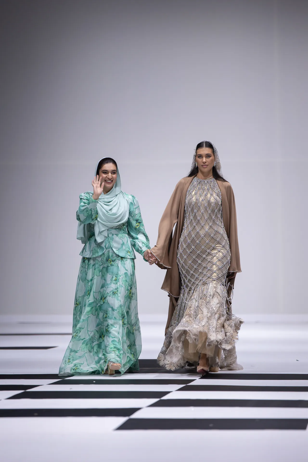 Two models hold hands and walk a fashion runway; left model wears a mint green floral hijab outfit, waving, right model wears a beige, beaded gown with a cape.