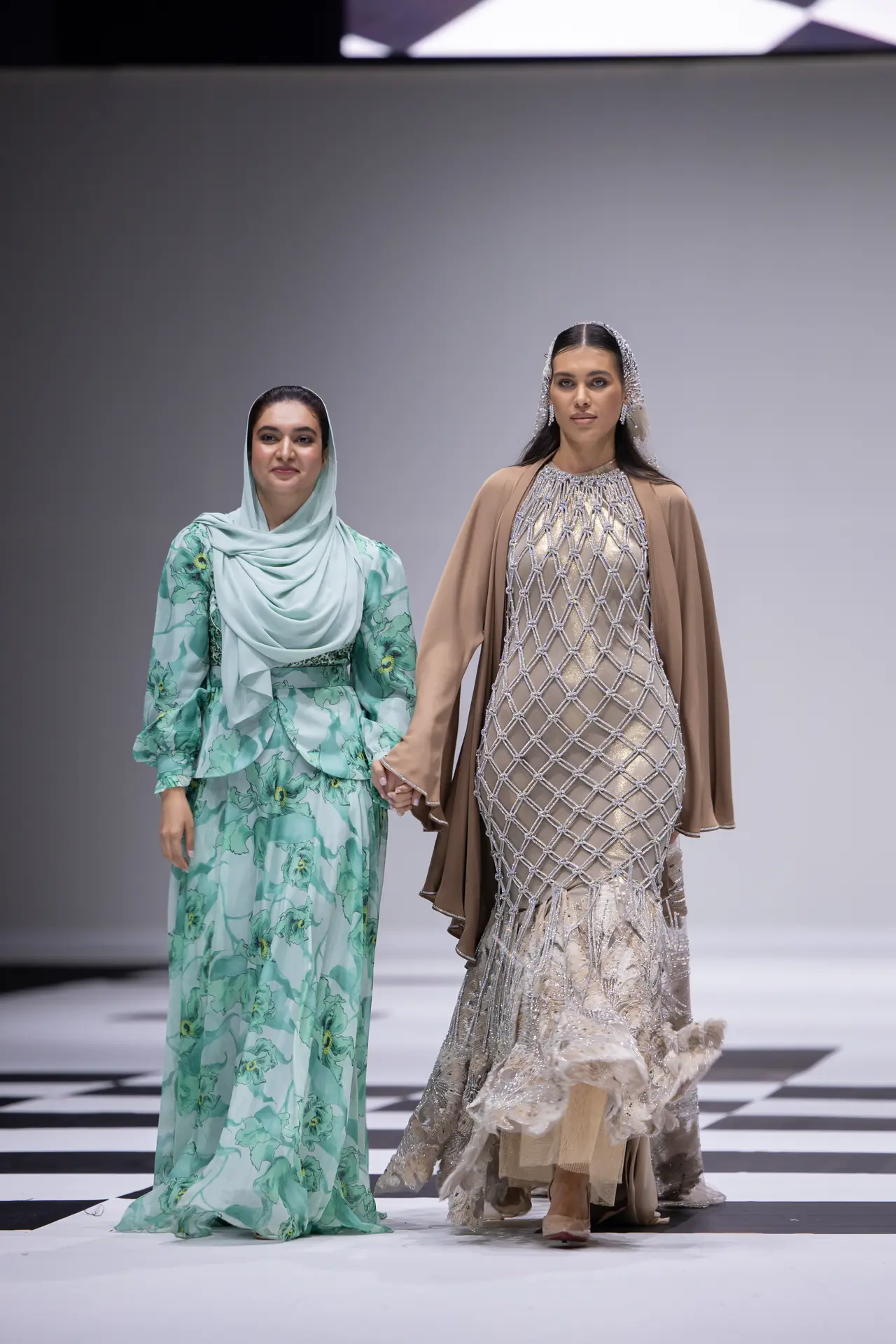 Two models walk hand in hand on a fashion runway; the left wears a mint green floral dress with a light hijab, the right a beige beaded gown with a cape.