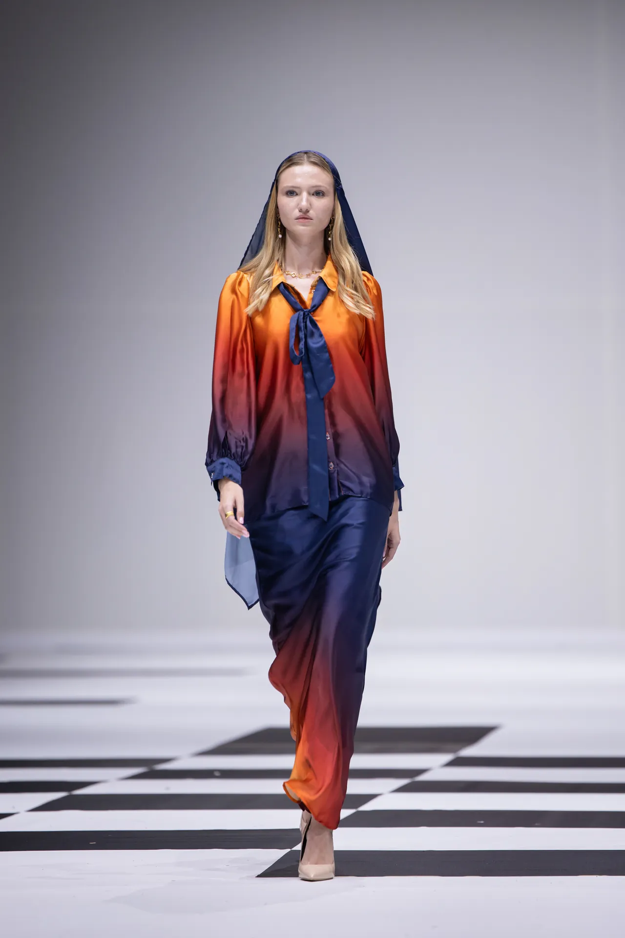 Model walking a fashion runway in a gradient orange-to-navy blouse and matching pants, with a navy headscarf and nude heels.