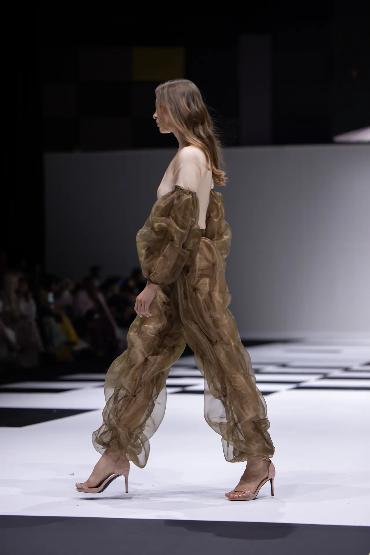 Model walking a fashion runway in a sheer brown organza dress over a beige top, nude heels.