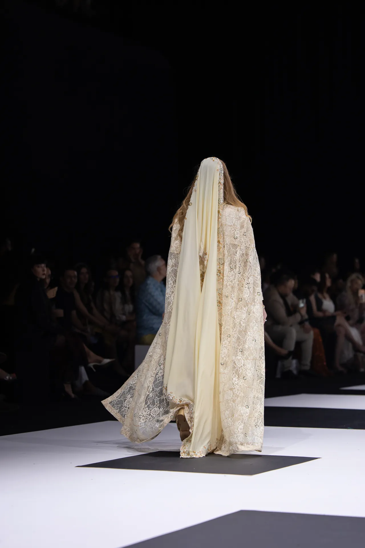 Back view of a model walking the runway in an ivory lace gown with a long cream veil, audience visible in the dark background.