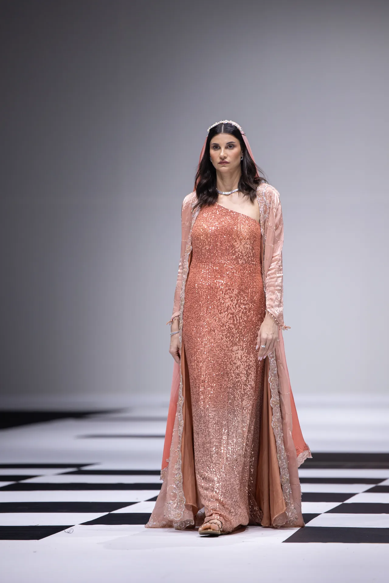 Model wearing a peach-pink sequined gown with a sheer matching cape on a black-and-white runway.