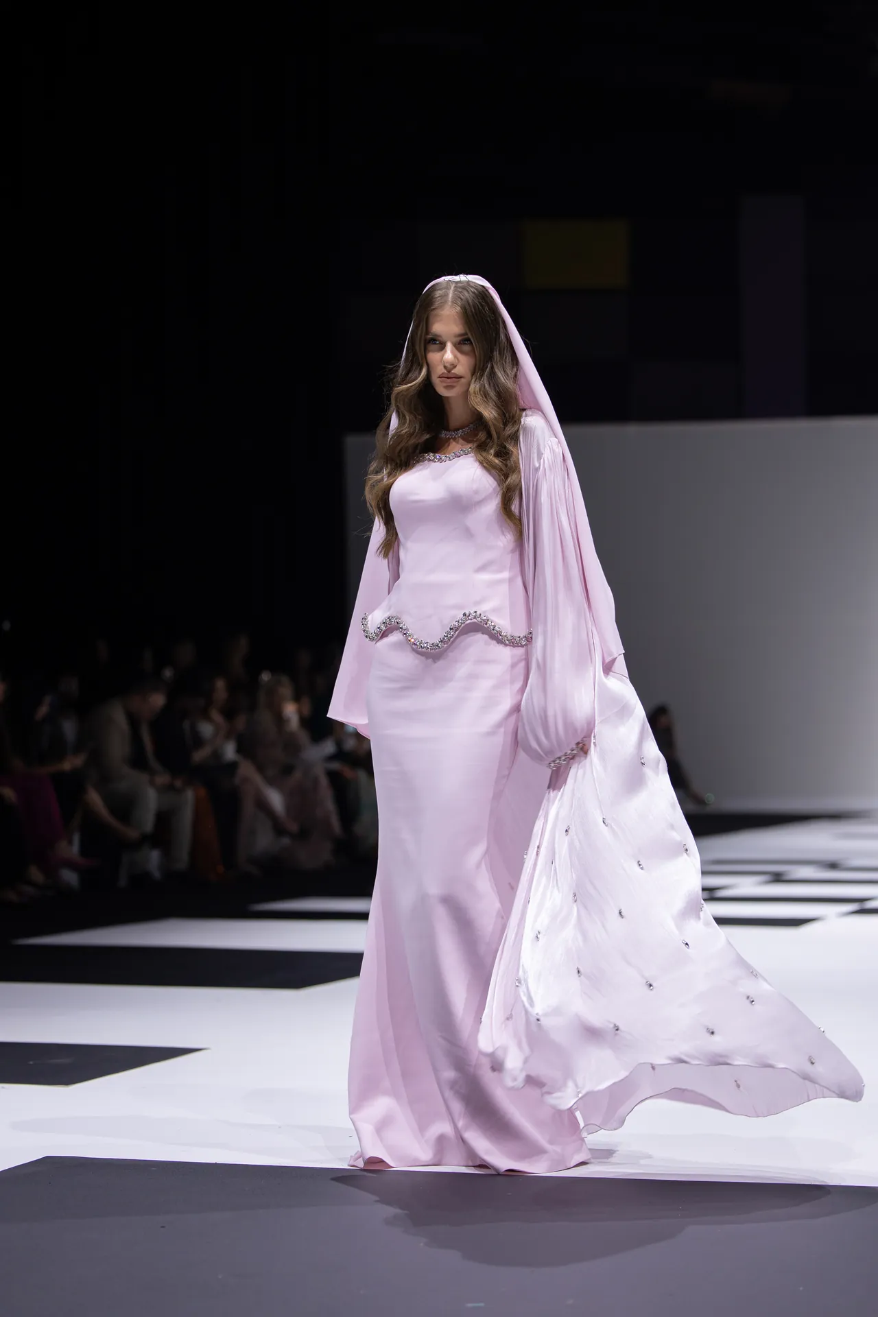 Model walks the runway in a pastel pink gown with a flowing cape and silver-trimmed detailing, under bright show lighting.