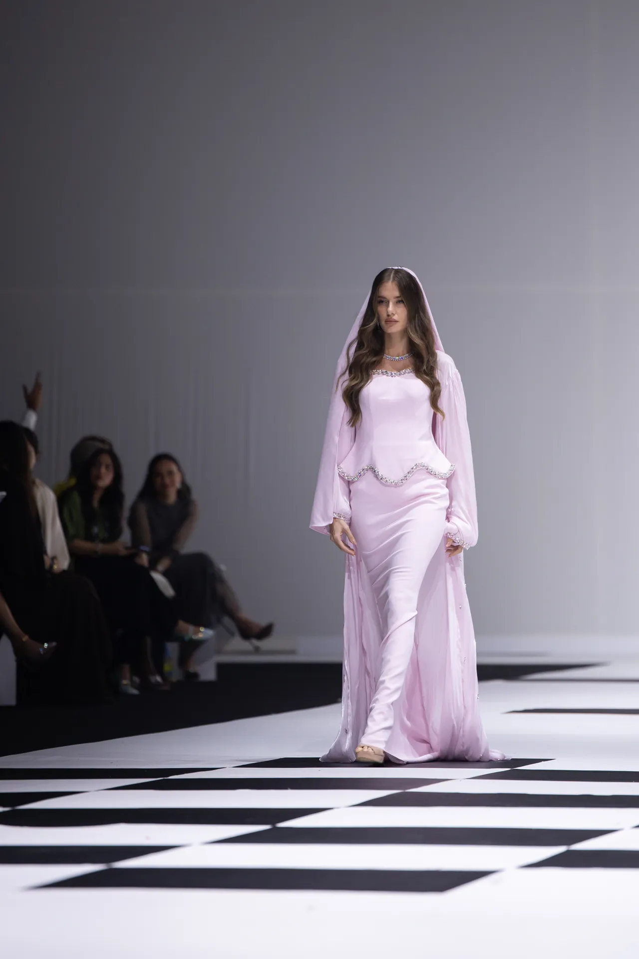 Model walking a pale pink gown down a black-and-white checkered runway with seated spectators on the left.