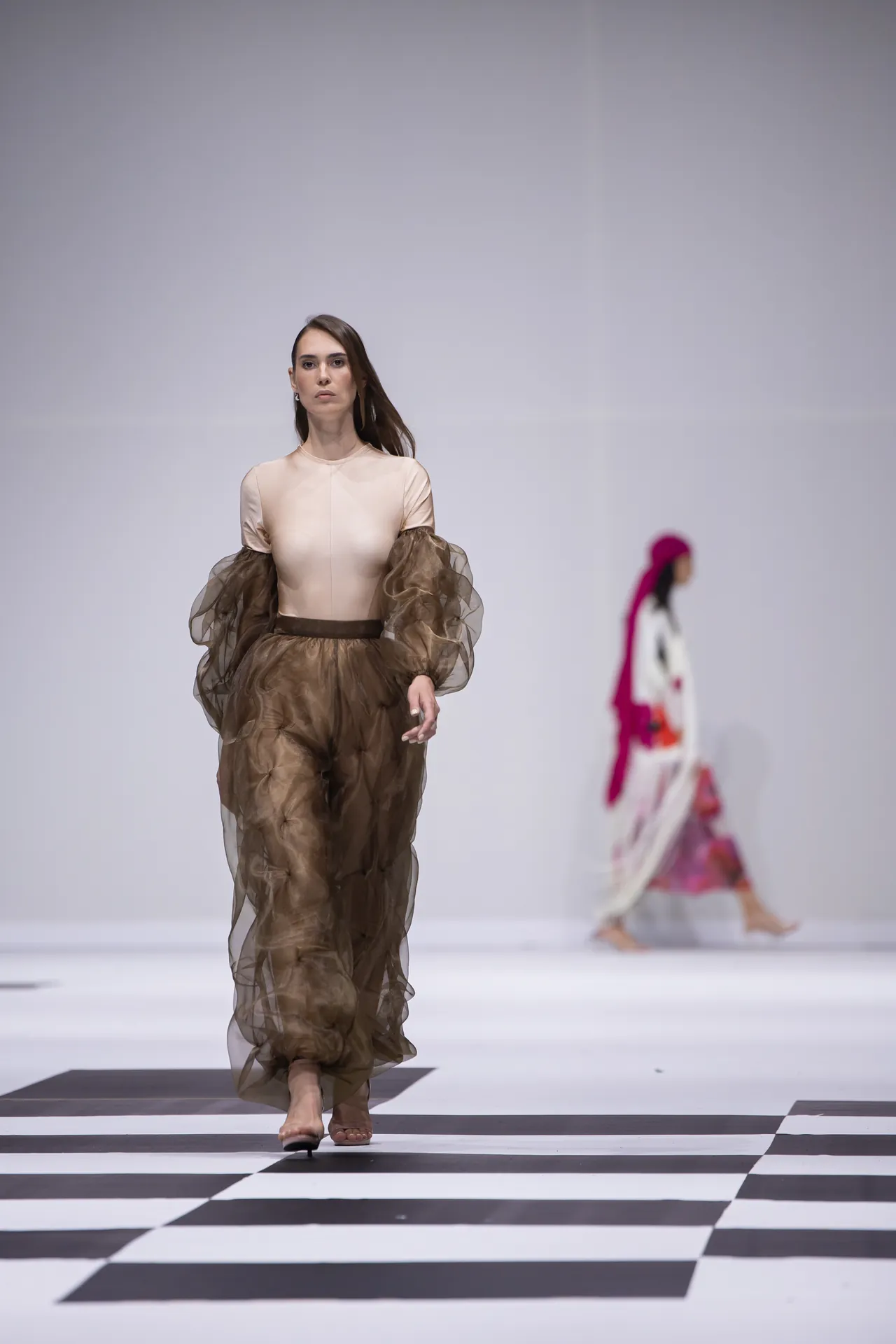 Model walks the runway in a beige top with voluminous sheer brown sleeves and matching sheer brown pants on a black-and-white checkered catwalk.