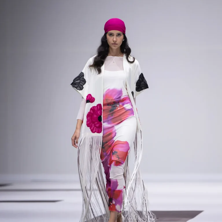 Model walking a runway in a white floral-print outfit with a hot-pink headscarf and long fringe cloak on a black-and-white checkered floor.