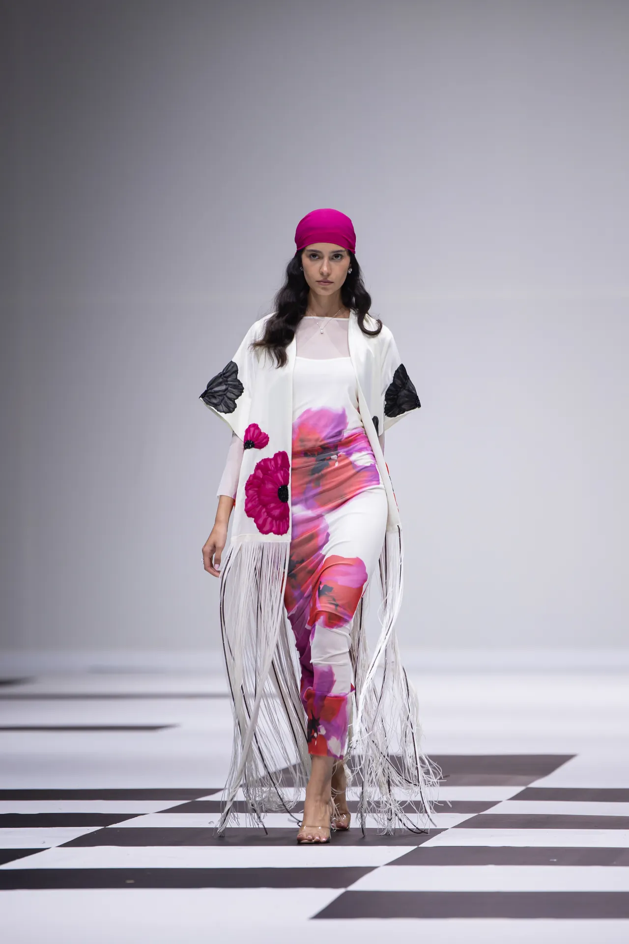 Model walking a runway in a white floral-print outfit with a hot-pink headscarf and long fringe cloak on a black-and-white checkered floor.