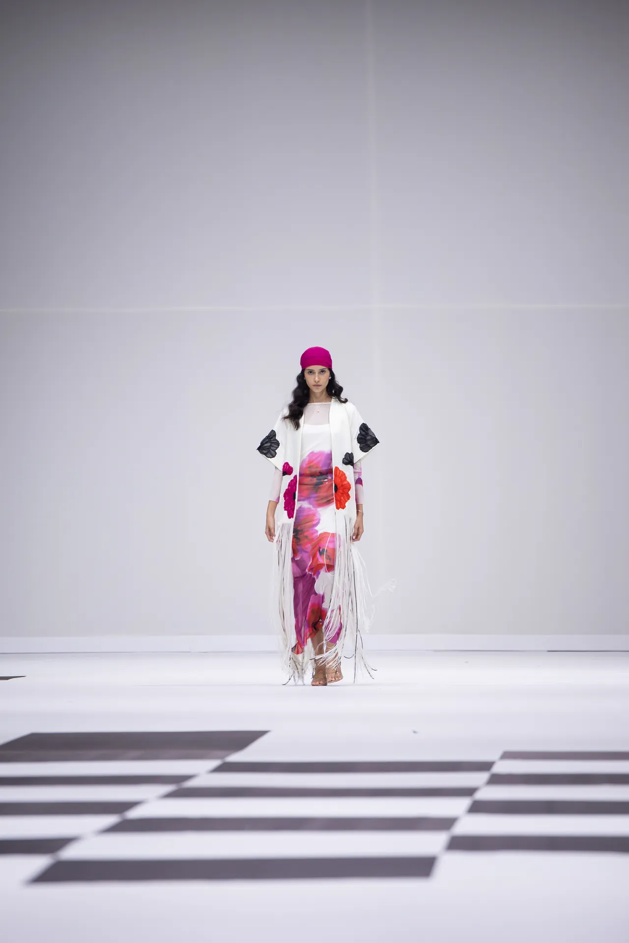 Model walking a fashion runway in a white floral-print dress with fringe and a pink headscarf against a minimalist backdrop.