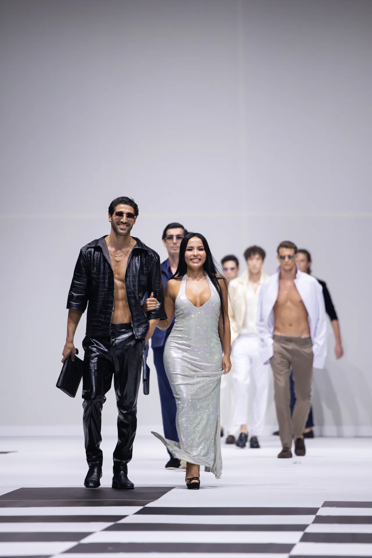 Models strut down a white runway in a fashion show; a man in black leather outfit leads beside a silver-dressed woman, with others in stylish outfits behind them on a striped floor.