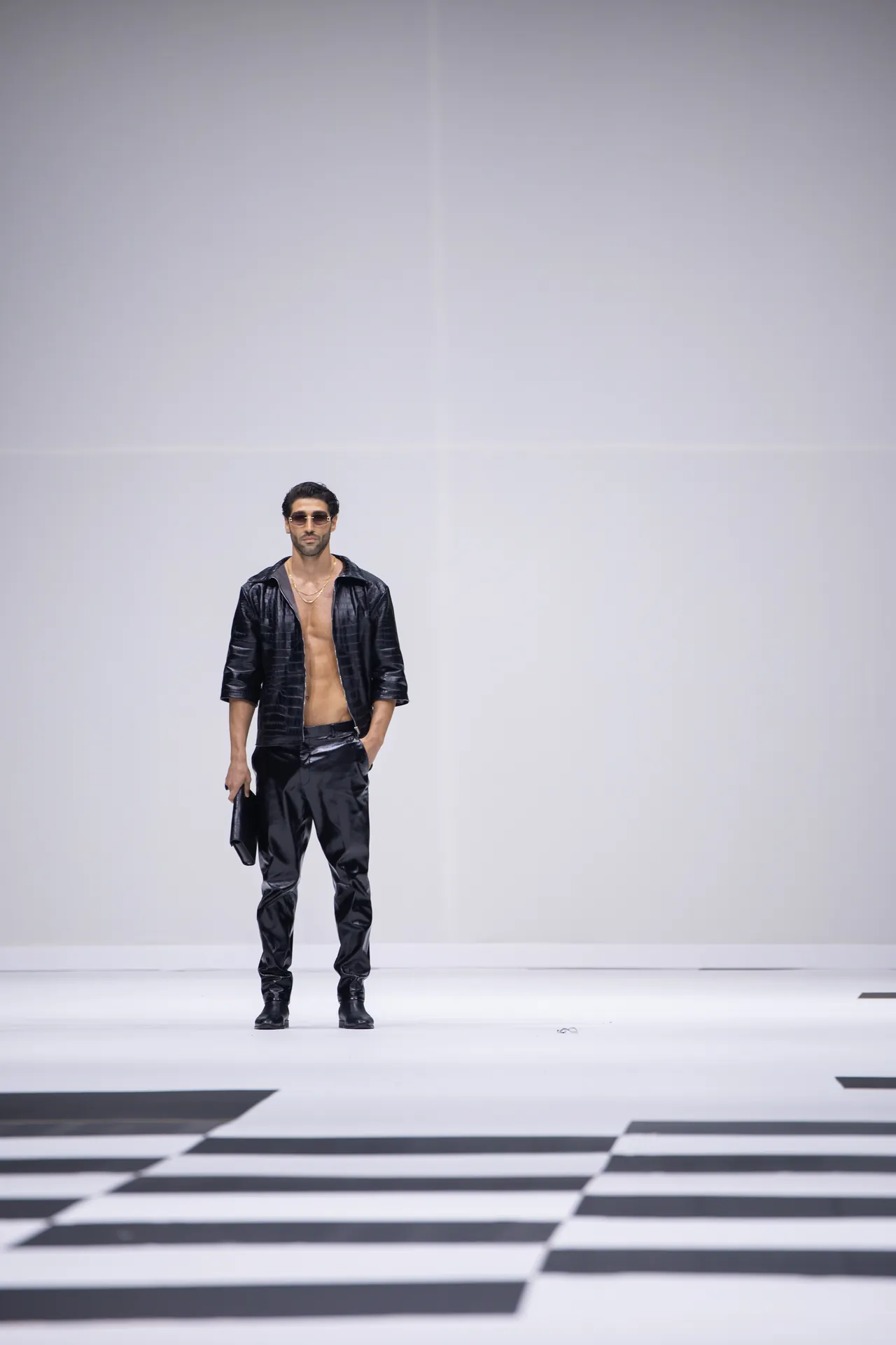 Male fashion model in a black shiny open jacket, no shirt, black pants, and sunglasses standing on a white runway with a black-and-white checkered floor pattern.