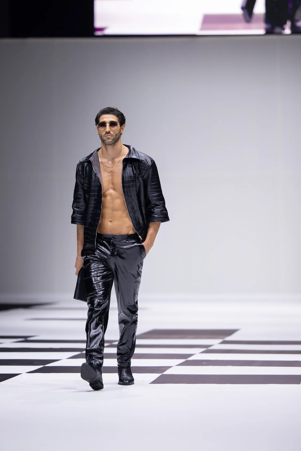 Male model walking the runway in a black shiny leather jacket and pants, chest exposed, with gold chains and sunglasses.