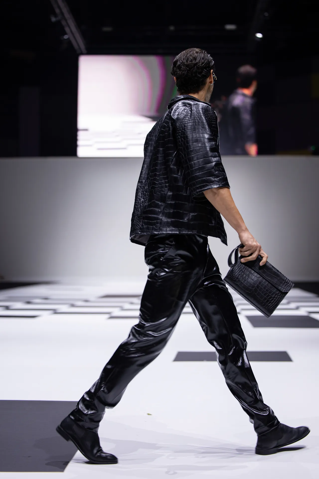 Model walking a fashion show runway in a glossy black crocodile-textured outfit, holding a matching black bag.
