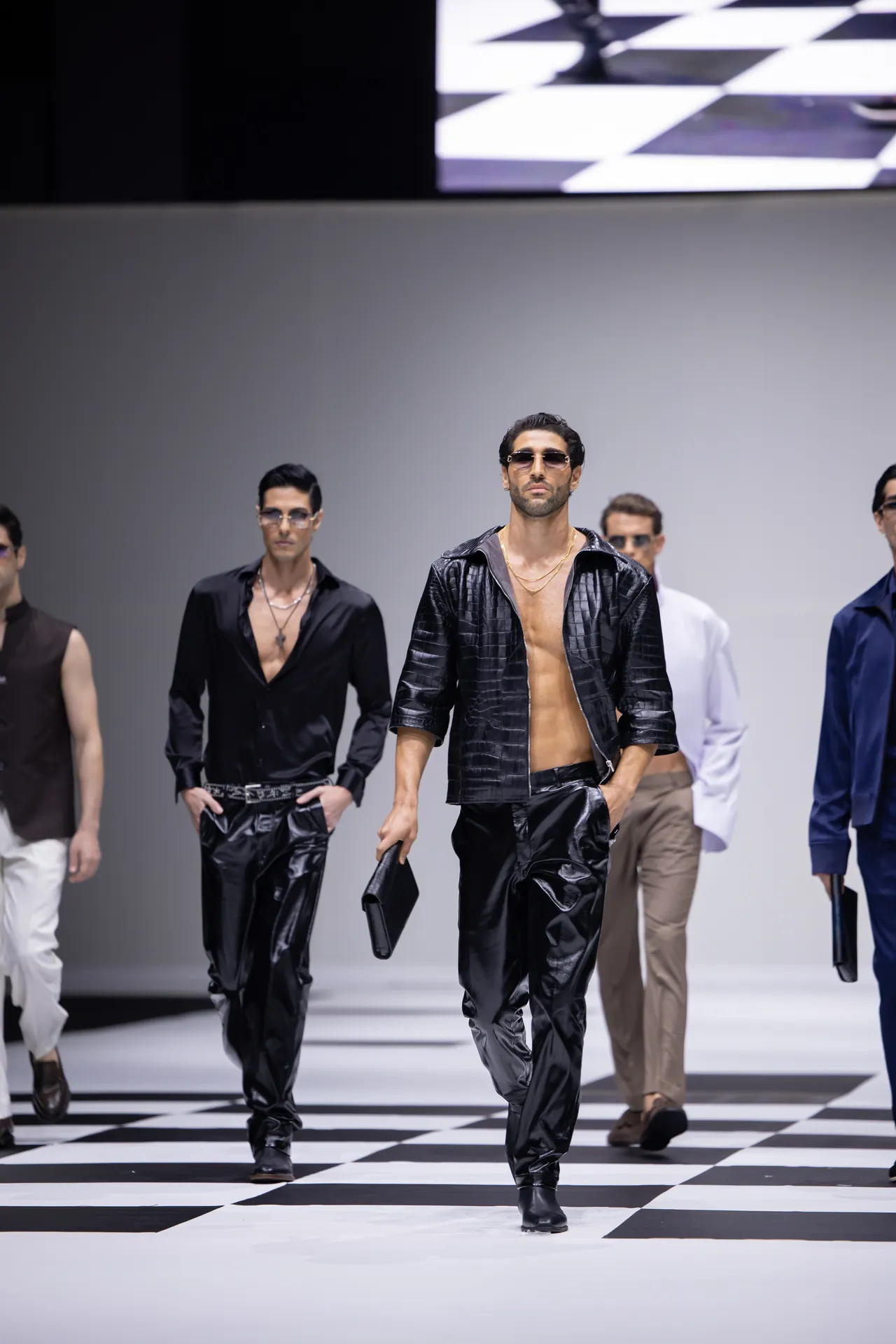 Male models walk a fashion runway in glossy black outfits on a black-and-white checkerboard floor; central model wears an open leather jacket, sunglasses, and gold chains, carrying a small bag.
