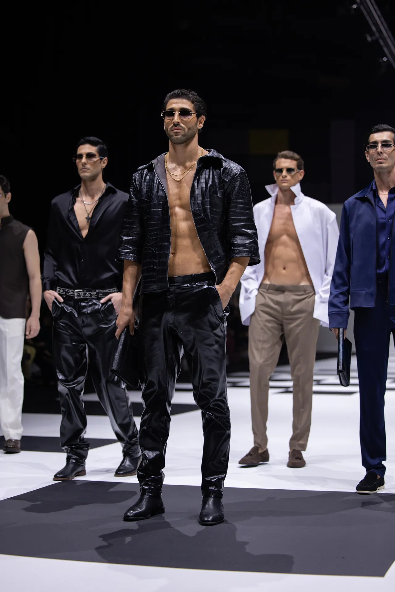 Male model on a fashion runway wearing a shiny black leather-look jacket, open chest with gold chains and sunglasses, with other models in the background.