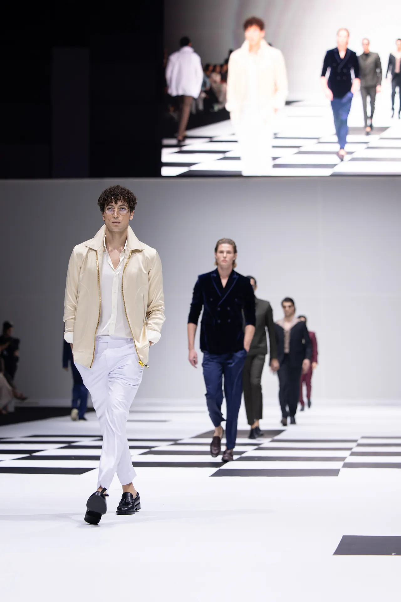 Male model in a beige jacket and white trousers walking a checkerboard runway, followed by other models in dark suits.