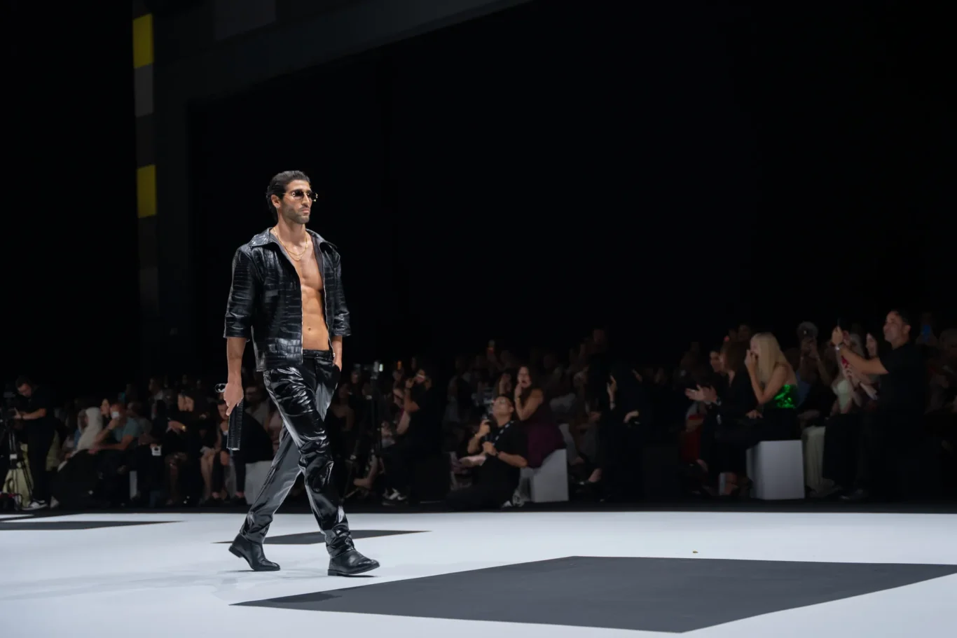 Male model walks a fashion show in a black leather outfit and sunglasses, audience watching from the dark background.