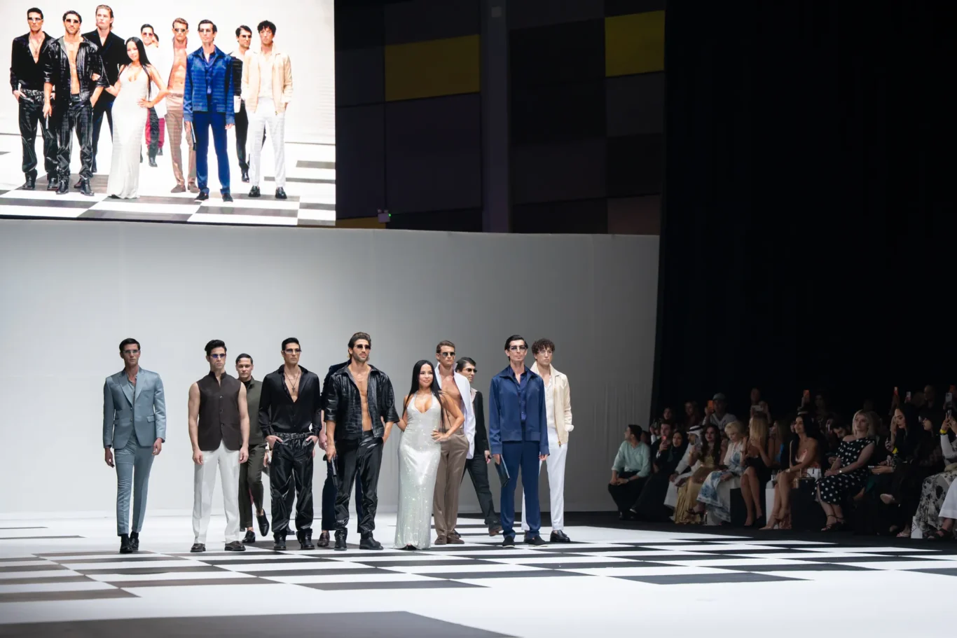 Group of models posing on a black-and-white checkered runway during a fashion show, audience on the right watching