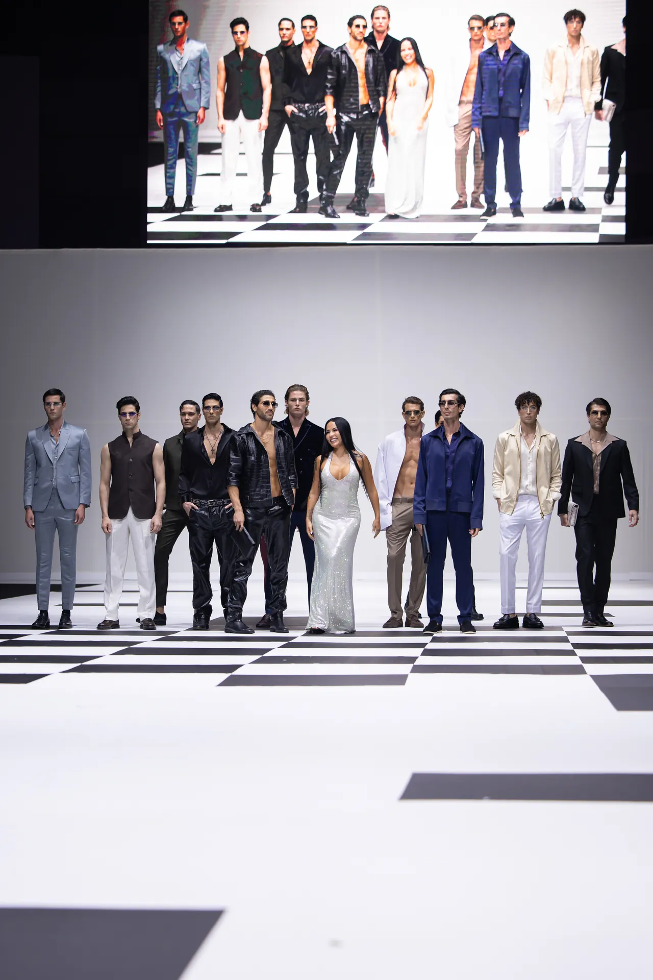 A line of models on a black-and-white checkerboard runway, wearing stylish suits and dresses, posing for a fashion show photo.