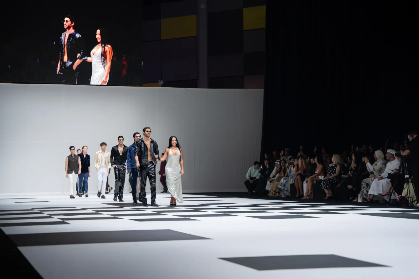 Models walk a fashion runway on a white checkerboard stage, audience seated along the right side watching eagerly.