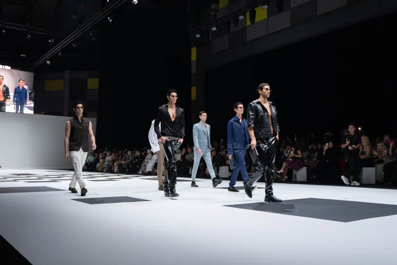 Male models walk a white runway in black and blue outfits at a fashion show, audience watching.