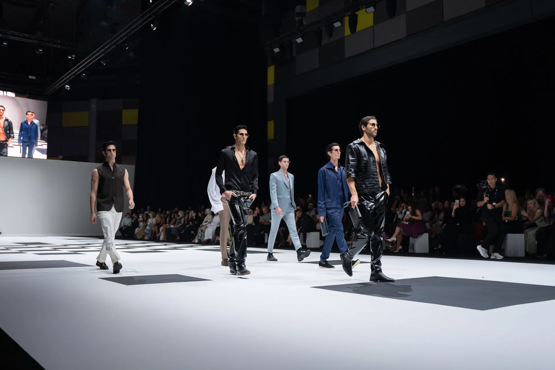 Male models walk a white runway in black and blue outfits at a fashion show, audience watching.