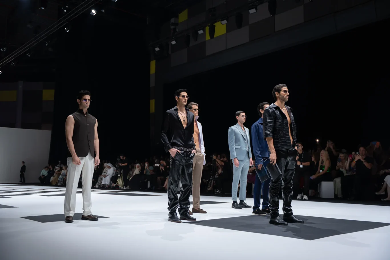 Male models walk the runway in stylish outfits during a fashion show, with an audience watching from the background