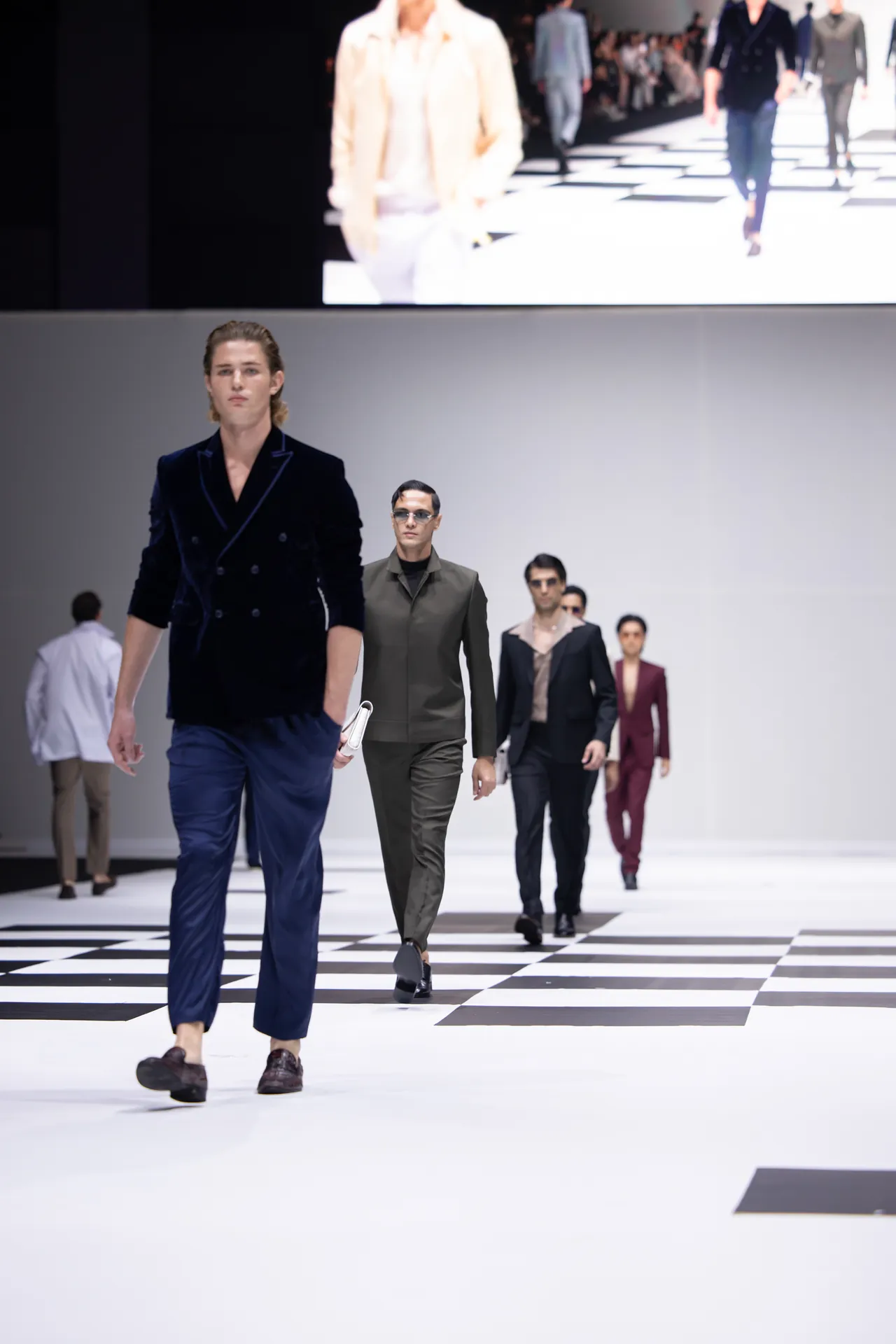 Models walk a black-and-white checkered runway in a fashion show, wearing tailored dark suits and sunglasses in a bright, minimalist venue.