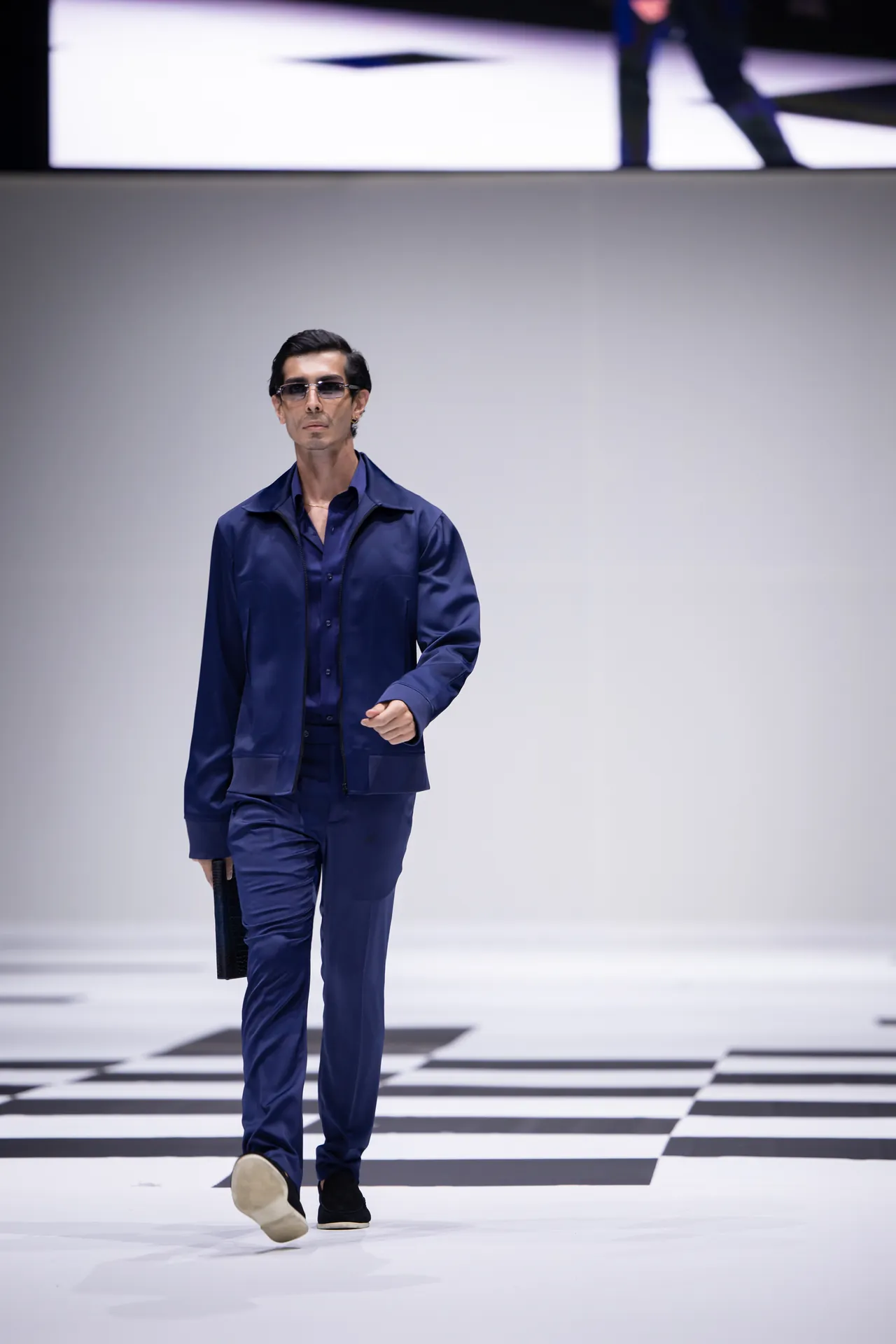 Male model walking a fashion runway in a navy blue suit and sunglasses.