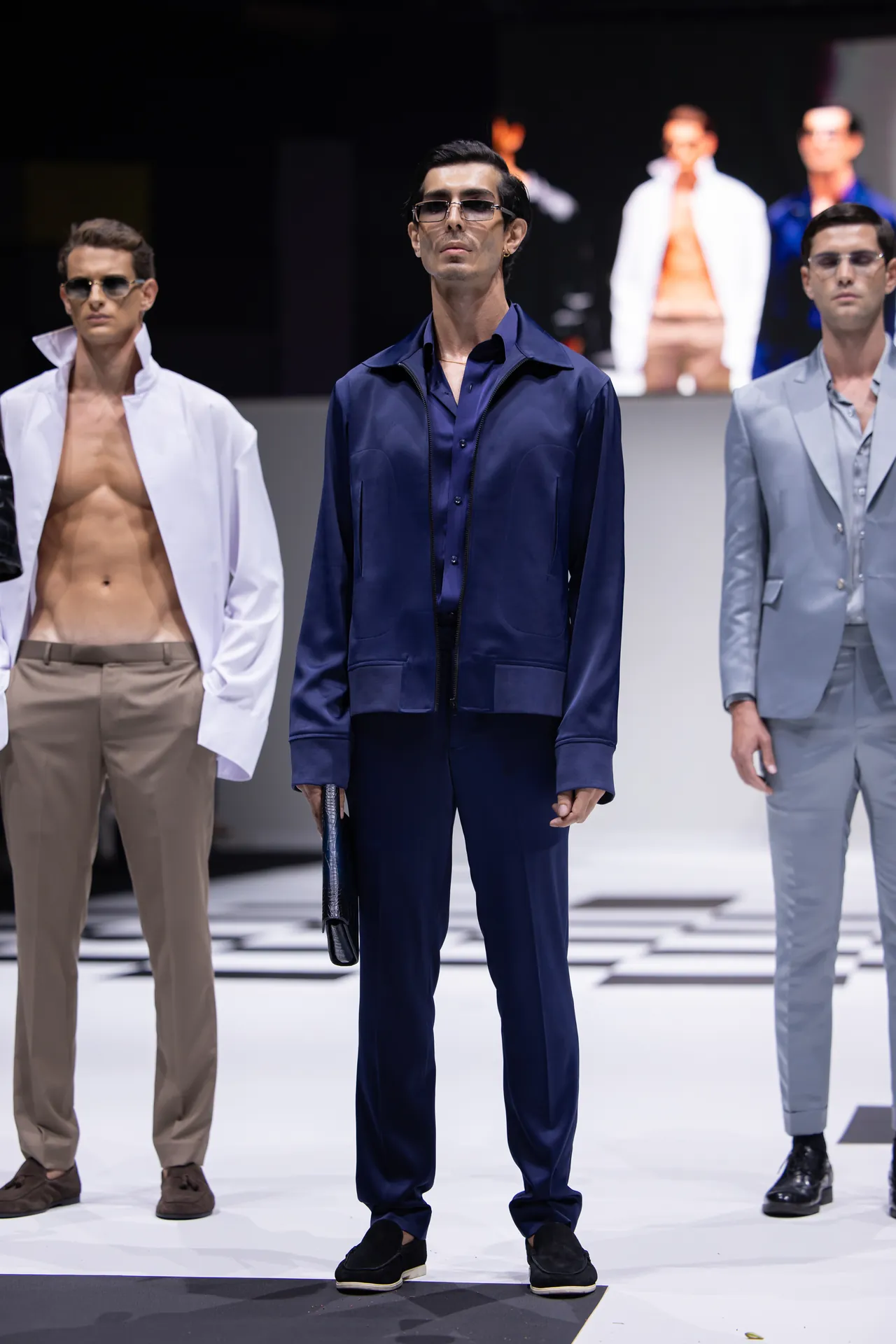 Male model in a navy blue suit and sunglasses standing on a fashion runway, flanked by two other models in beige and gray suits.