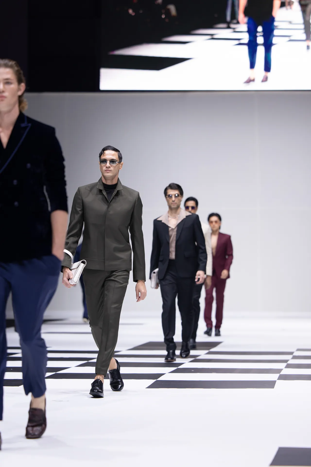 Model in an olive-green suit walks down a black-and-white checkered runway, other models follow in the background.