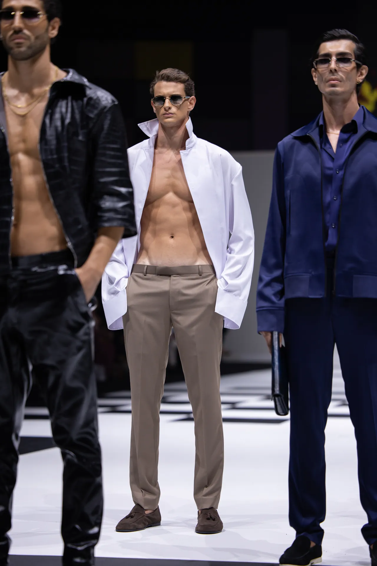 Male model wearing an open white shirt, beige trousers, and sunglasses on a fashion runway.