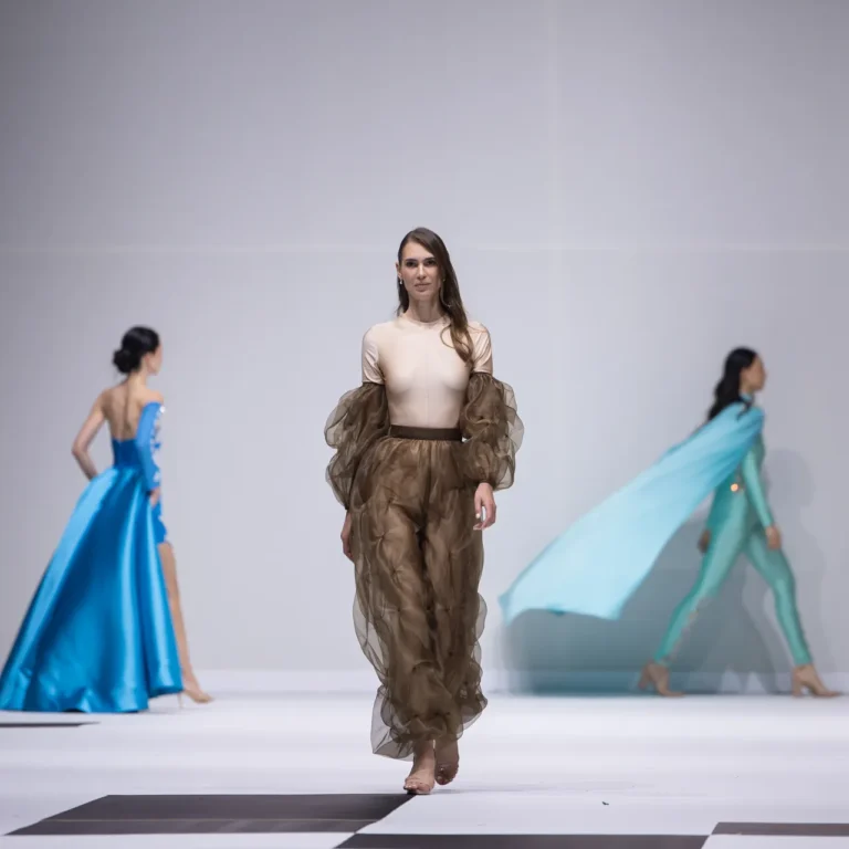 Model walks the runway in a beige top and brown sheer voluminous pants; blue and turquoise gowns are visible on either side of the catwalk.