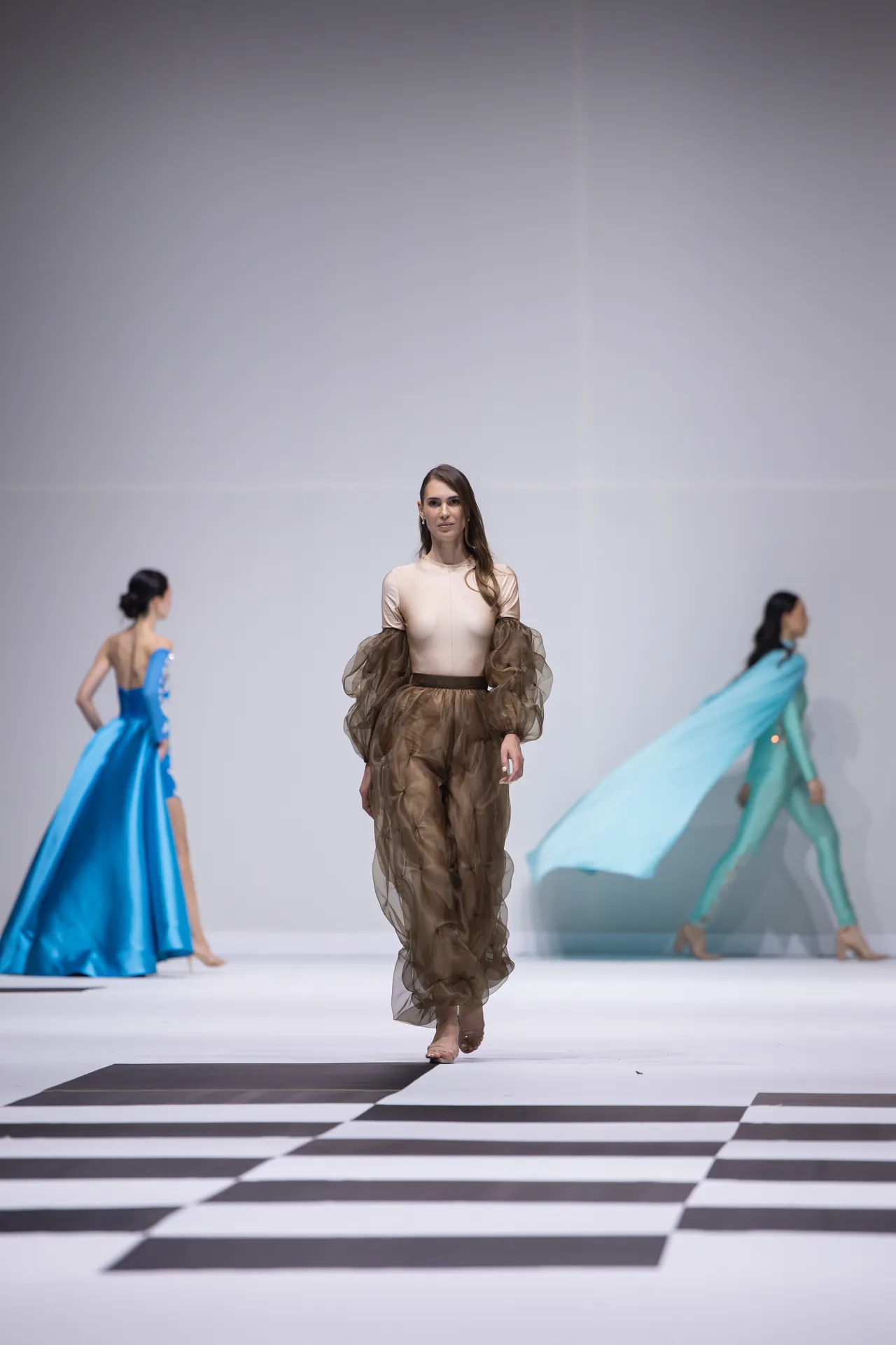 Model walks the runway in a beige top and brown sheer voluminous pants; blue and turquoise gowns are visible on either side of the catwalk.