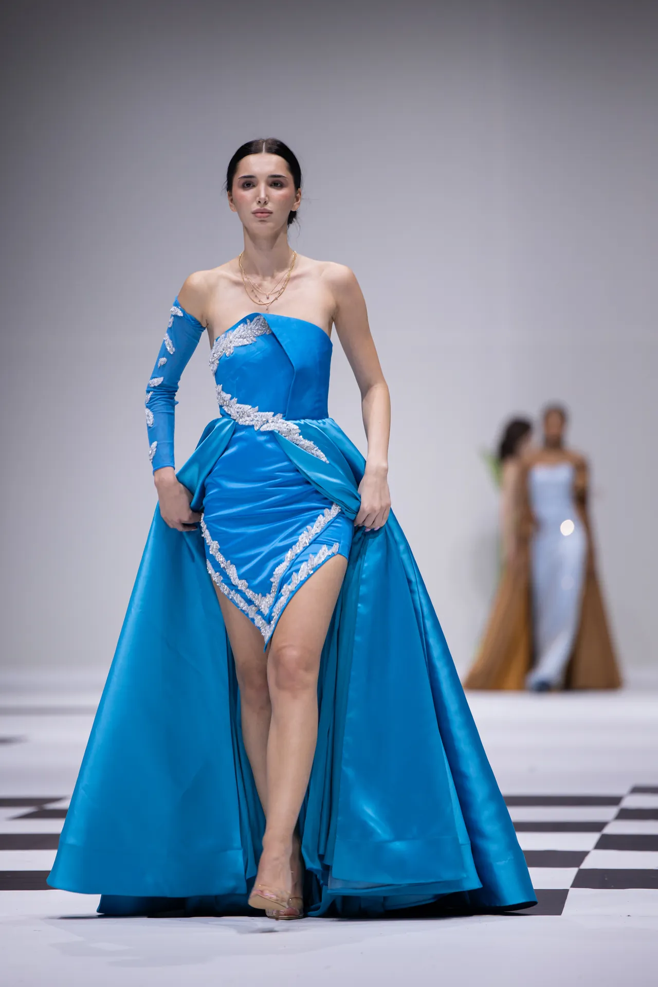 Model walking the runway in a vivid blue off-shoulder gown with silver embroidery and a full skirt on a checkered catwalk.