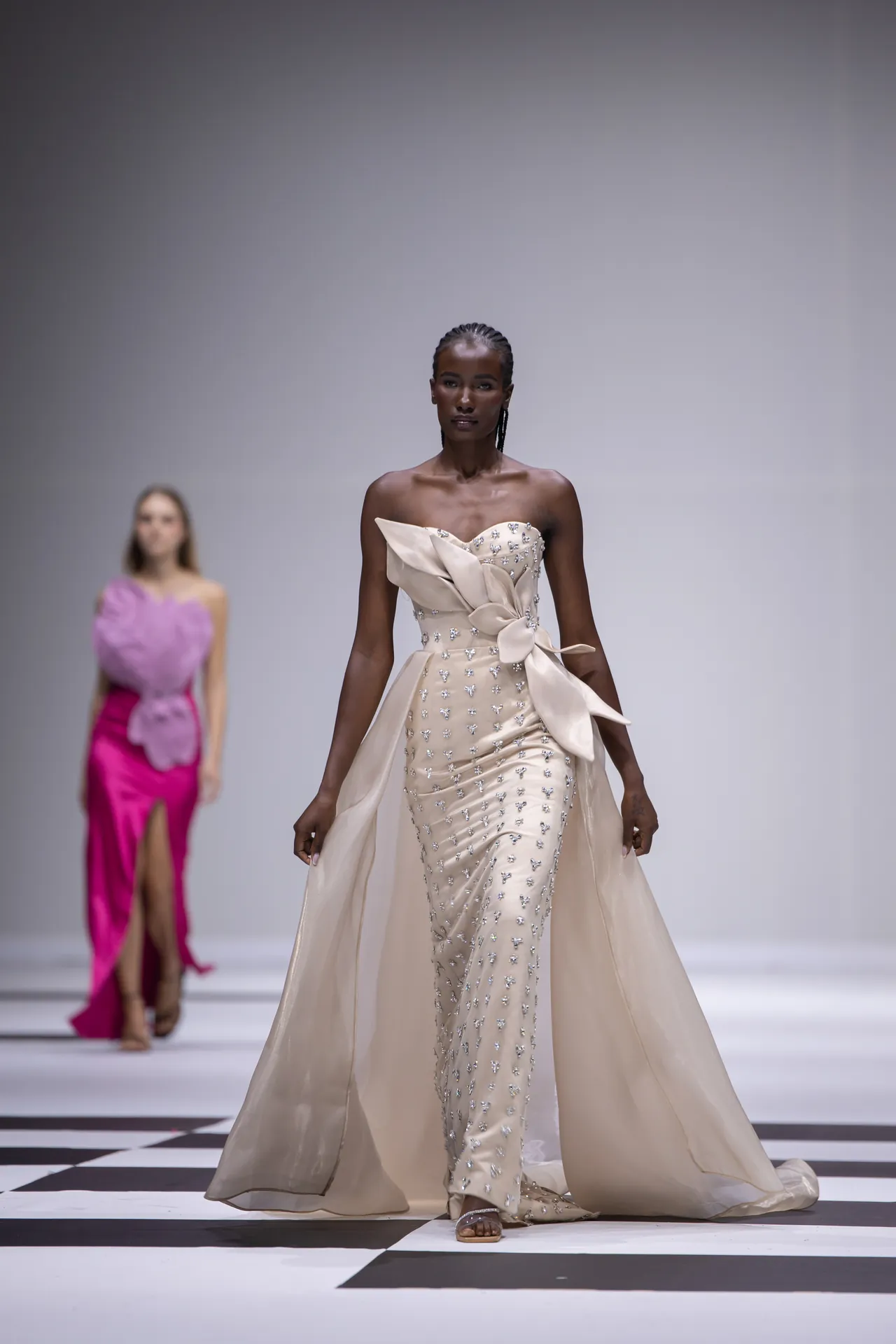 Runway model in a champagne strapless beaded gown with a large bow and flowing cape; another model in pink behind on a checkered catwalk.